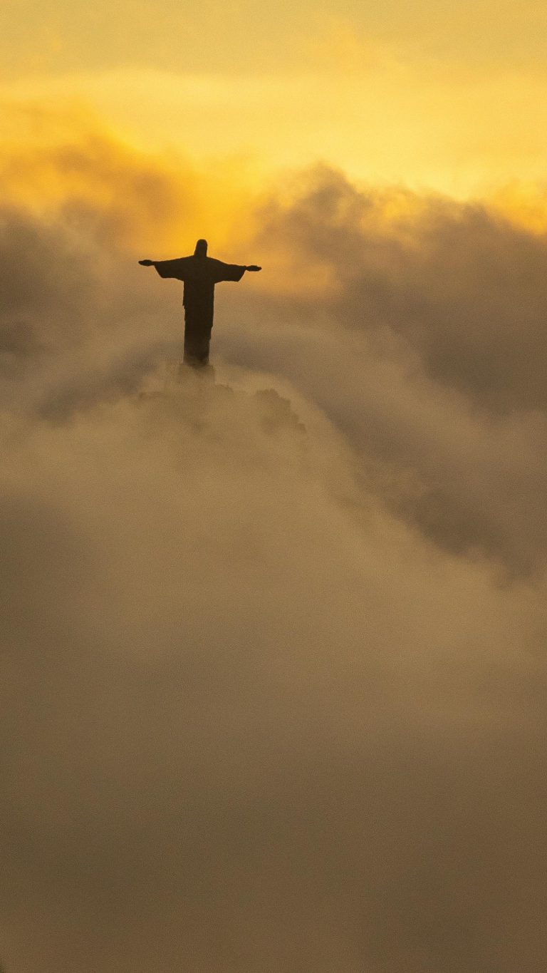 Silhouette of Christ the Redeemer statue emerging from clouds in Rio de Janeiro at sunrise.